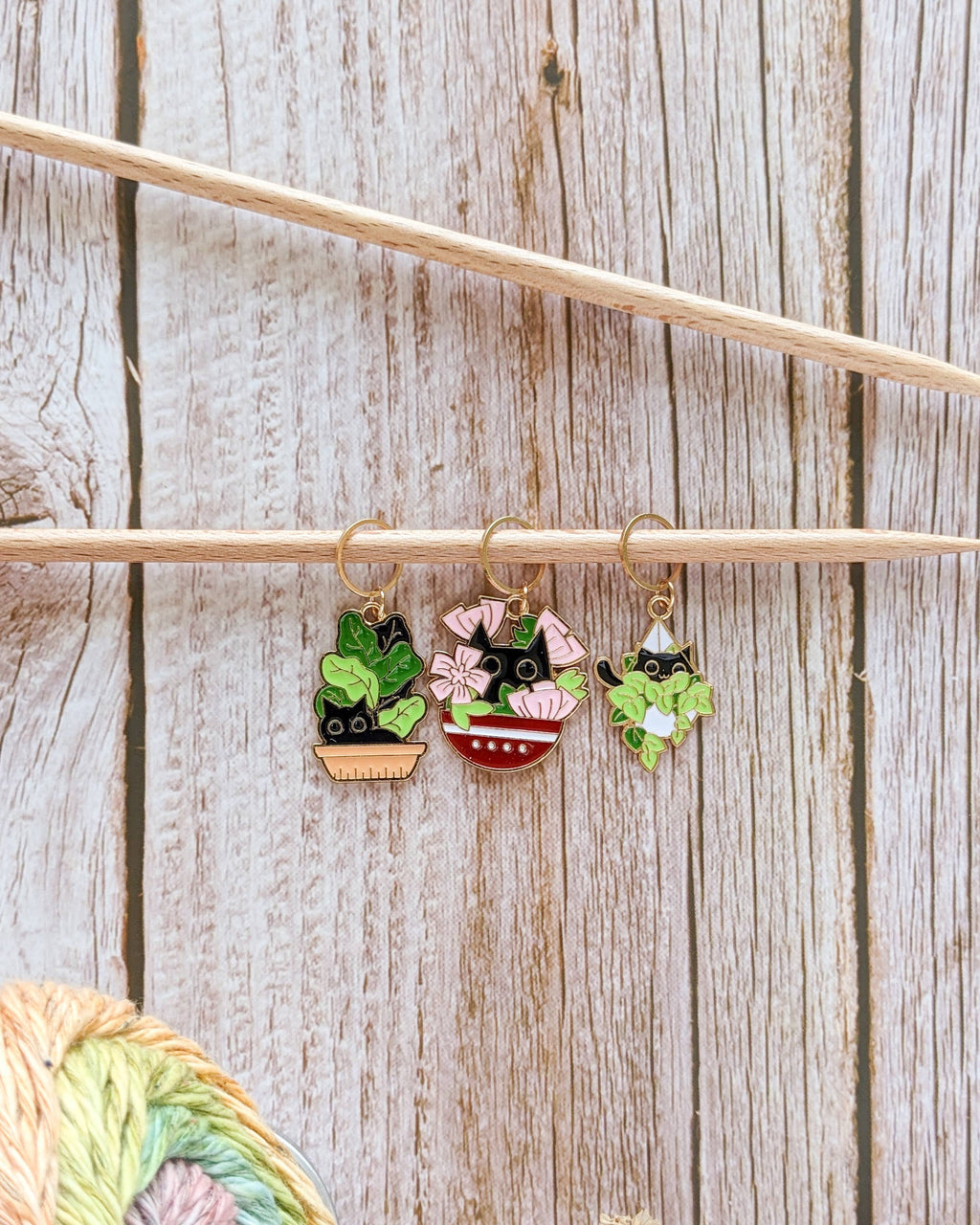 Three stitch markers with cats hiding in garden plant pots hanging from a knitting needle