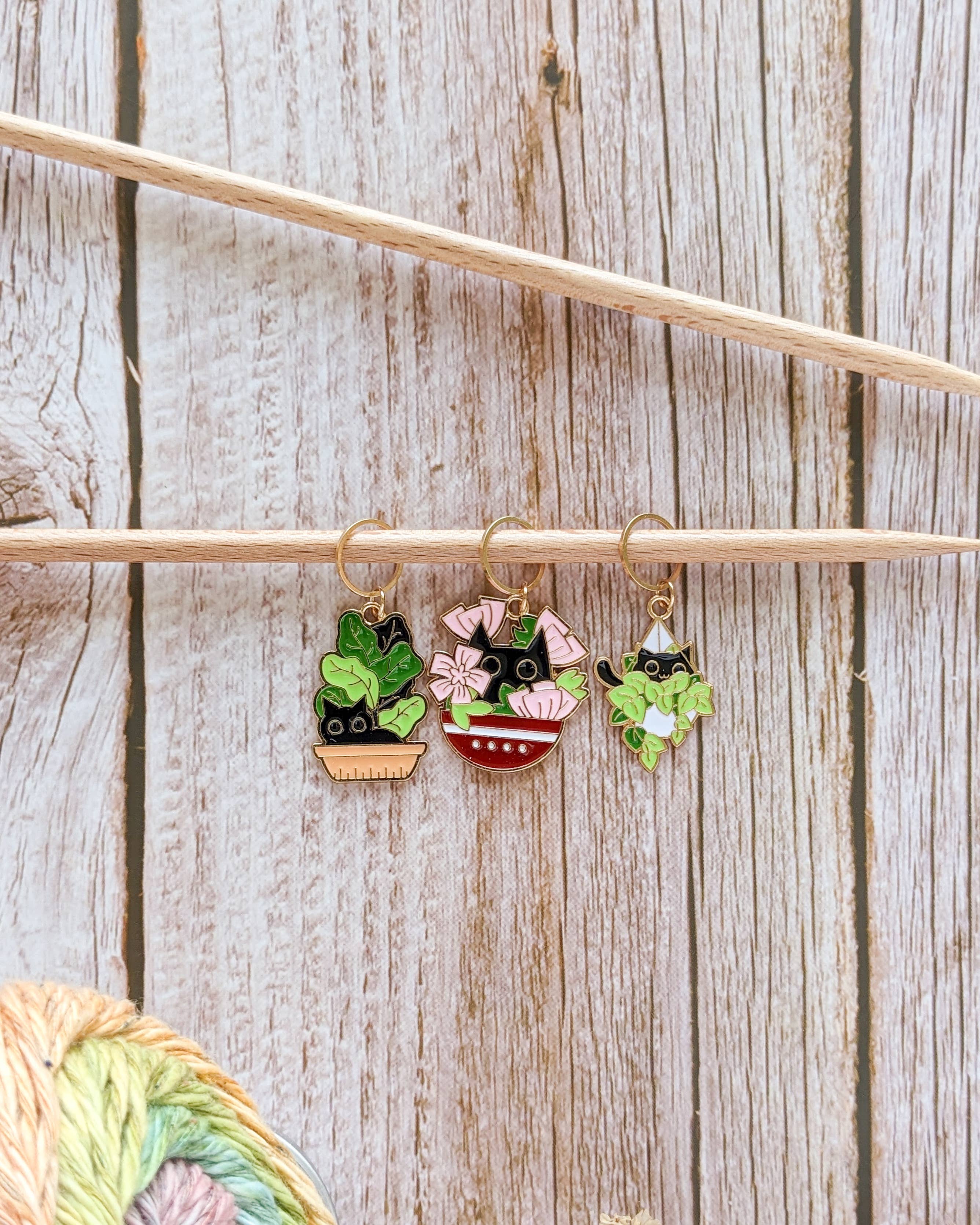 Three stitch markers with cats hiding in garden plant pots hanging from a knitting needle